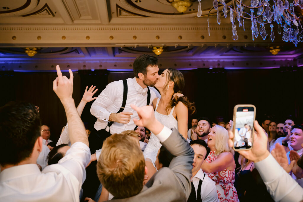 Bride and Groom being lifted in chairs by their guests as the other wedding guests look on and cheer.